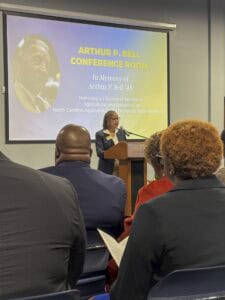 A speaker honors Arthur P. Bell during a dedication ceremony in a conference room at a university.