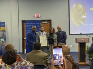 Four people pose together, holding a proclamation at an event, with an audience in the background.