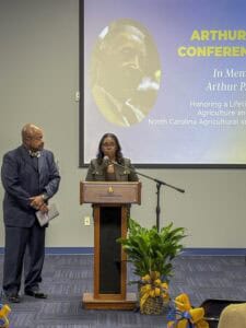A woman speaks at a podium while a man listens attentively at an event honoring Arthur P. in agriculture.