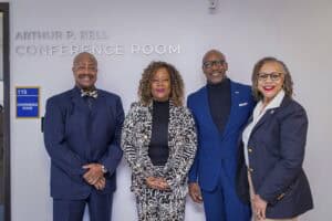 Four professionals pose together in front of a conference room sign, smiling and dressed in stylish suits.