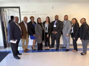 A group of nine professionally dressed individuals pose together in front of a wall with the sign "Arthur P. Bell Conference Room." They are standing close together, smiling at the camera. The group includes men and women of diverse backgrounds, wearing business attire, including suits, blazers, and dresses. One man on the far left holds a document in his hand. The setting is indoors with a bright hallway and an open door in the background.
