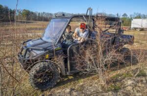 A man wearing sunglasses, a gray shirt, and an orange cap sits in the passenger seat of a black all-terrain utility vehicle, leaning out to examine a small tree branch. A young boy is in the driver’s seat, looking at the controls. The vehicle is parked in a rural field with rows of leafless bushes, dry soil, and a clear blue sky. In the background, there are trees, a truck, and other farm equipment.