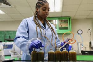 A female scientist wearing a blue lab coat and purple gloves works in a laboratory. She has long braided hair and is using a device with tubes to analyze liquid samples in multiple glass bottles filled with dark-colored fluids. The laboratory features green cabinets, a chalkboard with chemical formulas, and various scientific equipment in the background.
