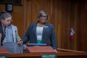 Councilperson Jamilla Pinder stands confidently in a wood-paneled council chamber, wearing a black blazer with a decorative brooch, a white blouse, and a green sash. She smiles while holding onto the back of a chair, with two individuals nearby engaged in conversation. A nameplate in front of her reads "Jamilla Pinder, Councilperson - At Large."