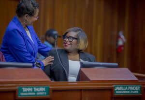 A woman in a royal blue blazer stands and places her hand on the shoulder of a seated councilperson wearing a black blazer and glasses. They smile warmly at each other inside a wood-paneled council chamber. Nameplates in front of them read "Jamilla Pinder, Councilperson - At Large" and "Tanni Thurn, Councilperson - District 5."