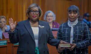 A woman wearing glasses and a dark green blazer raises her right hand while taking an oath of office. She has a confident expression and is standing in a formal setting with wooden paneling. Another woman, dressed in a black and white checkered jacket over a white turtleneck, holds a book with a serious expression. Behind them, individuals are seated, including one wearing a purple blazer, observing the ceremony.