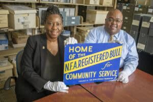 Two individuals hold a vintage sign about community service, surrounded by archival boxes in a storage area.