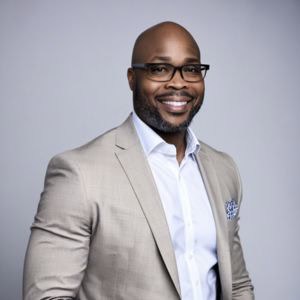 A professionally dressed man with a neatly trimmed beard and glasses smiles confidently at the camera. He is wearing a light beige suit jacket over a white dress shirt, complemented by a patterned pocket square. The background is a solid, neutral gray.