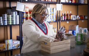 A food pantry staff member wearing glasses, a cream-colored sweater with red trim, and a denim shirt sorts through a wooden crate filled with snack bars and other food items. She appears to be organizing or distributing supplies. Behind her, shelves stocked with canned goods, boxed foods, and bottles of cooking ingredients line the pantry wall.