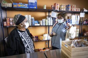 A food pantry staff member in a denim shirt and face mask holds up a can of food while speaking with a visitor wearing a black coat and headband. The visitor listens attentively, holding a red phone case. Behind them, shelves are stocked with canned goods, pasta, and other pantry staples, with small signs labeling different sections.
