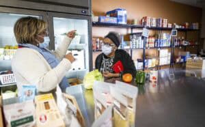 A staff member and a visitor, both wearing face masks, converse inside a food pantry. The staff member, wearing a light-colored sweater and blue scarf, gestures toward a refrigerator filled with fresh produce. The visitor, dressed in a black coat and headband, listens attentively with a red phone case in hand. Shelves stocked with canned goods, dry foods, and other pantry staples line the background.