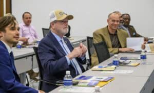 A group of professionally dressed individuals sit at long tables in a conference-style room, engaged in discussion. A man in a navy blue suit, wearing a beige baseball cap with a logo, sits in the foreground, appearing attentive. To his left, a younger man in a blue suit looks toward the front of the room. Other participants, including a man in a tan blazer smiling, are seated behind them, some holding documents or taking notes. The tables have bottled water, brochures, pens, and notepads neatly arranged.