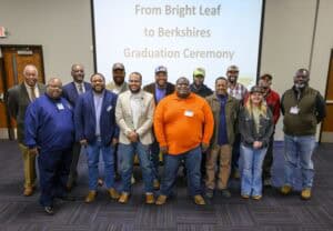 Group of fifteen people posing for a photo in front of a screen displaying the text 'From Bright Leaf to Berkshires Graduation Ceremony at North Carolina Agricultural and Technical State University'