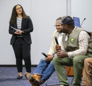 A woman in a black blazer and striped shirt stands in a room with blue carpet, holding papers and speaking. Two men sit on chairs in front of her, engaged in a discussion. The man on the left wears a light-colored suit jacket and jeans, holding a microphone. The man on the right wears a green vest over a plaid shirt, a cap, and green pants, also holding a microphone. A wooden podium and a computer monitor are in the background.