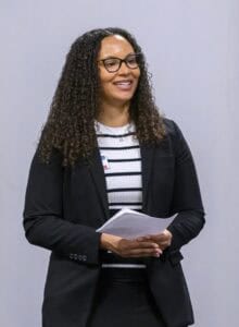A woman with curly hair stands in a room, holding papers in her hands. She wears a black blazer over a white shirt with black horizontal stripes and glasses.