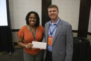 A woman wearing glasses and an orange top smiles while holding an envelope, standing next to a man in a grey blazer who is also smiling. The man wears a name tag that reads "NC PORK". They are posing indoors.