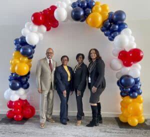 Four professionally dressed individuals stand side by side, smiling at the camera under a colorful balloon arch. The arch is made of red, white, blue, and yellow balloons. The group consists of one man in a beige suit with glasses and three women in dark business attire, one of whom is wearing a yellow blouse. The background features a plain white wall and a carpeted floor.