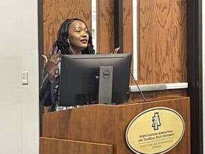 A woman with dark, wavy hair and a confident expression stands behind a wooden podium, gesturing with her hands while speaking. She is wearing a striped blazer and has neatly manicured nails. A Dell computer monitor and a microphone are positioned in front of her. The podium bears the logo and name of North Carolina Agricultural and Technical State University, specifically the Agriculture and Environmental Sciences department. The background features wooden paneling and a partially visible window or door.