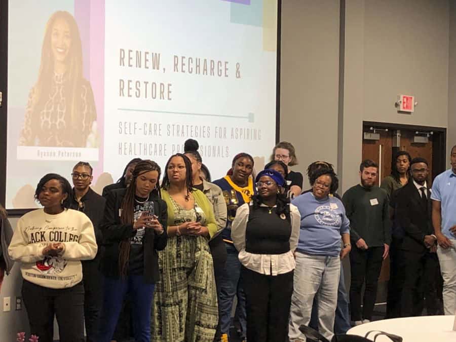 A diverse group of people stands in a conference room in front of a large projector screen displaying a presentation titled "Renew, Recharge & Restore: Self-Care Strategies for Aspiring Healthcare Professionals" by Ryanne Peterson. The attendees, dressed in casual and semi-formal attire, appear engaged, with some looking at their phones, clasping their hands, or listening attentively. The setting includes a neutral-colored wall, double doors, and a visible exit sign in the background.