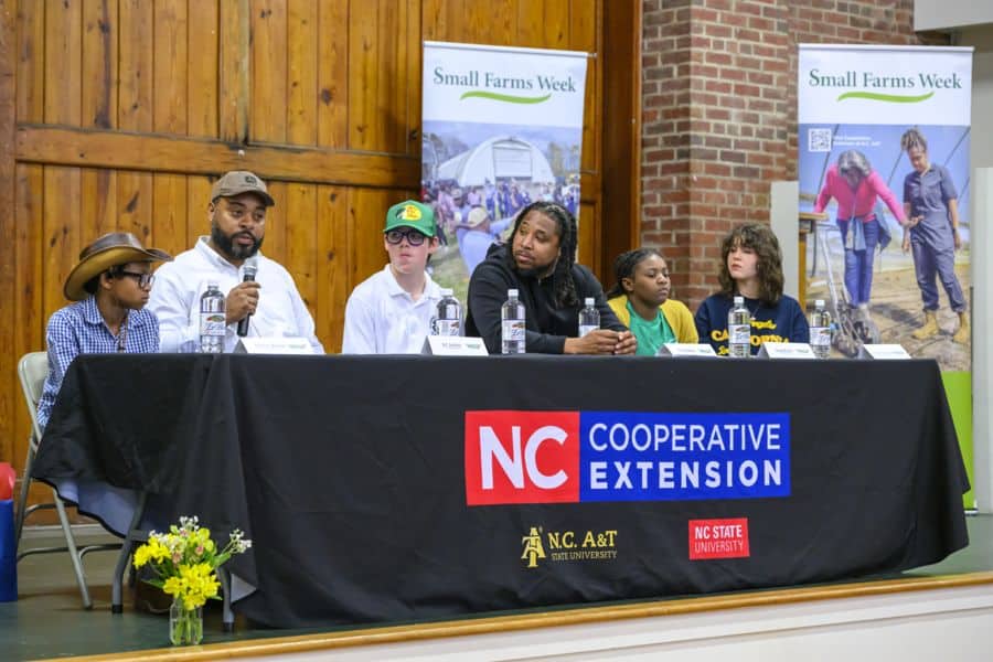 A panel of six individuals sits behind a table with an NC Cooperative Extension banner at a Small Farms Week event. One man speaks into a microphone while others listen. The panel includes youth and adults of diverse backgrounds. Behind them are banners promoting Small Farms Week, with images of farming activities. A vase of yellow flowers sits in front of the table.