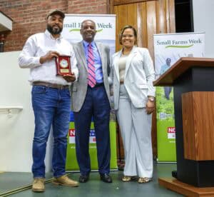 Three individuals stand smiling at a Small Farms Week event. The man on the left holds a wooden plaque with a gold-rimmed clock. Behind them are promotional banners for Small Farms Week, NC Cooperative Extension, and NC State University. They are positioned in front of a wooden podium and brick wall.