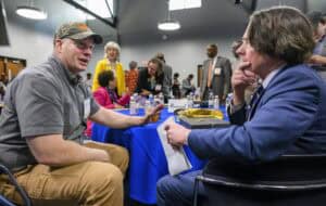 Two men sit and talk at a conference table. One wears a camouflage cap, while the other is in a suit holding a notepad. People in the background engage in conversations. Various materials and water bottles are on the table.