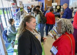 A lively networking event with several people interacting in a bright room. Two women are engaged in conversation in the foreground. Tables with informational displays line the walls, and attendees are browsing the exhibits.
