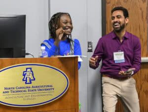 A young woman speaks into a microphone at a podium labeled North Carolina Agricultural and Technical State University, while a man beside her smiles and claps. Both appear engaged and happy.