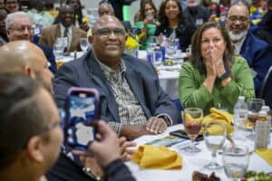 Man and woman seated at a banquet table, reacting with joy and surprise, surrounded by smiling attendees; the woman covers her mouth with her hands while the man smiles warmly, as someone in the foreground captures the moment on a phone.