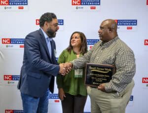 Three people are standing in front of a white backdrop with NC Cooperative Extension logos. Two men are shaking hands; one holds a plaque reading Small Farms Week, while a woman stands between them, smiling.