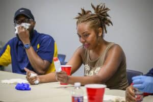 A woman holding an ice pop and smiling sits at a table with red cups, while a man beside her wipes his mouth with a napkin. Both appear to be participating in an indoor activity or event.