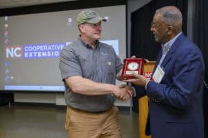 Two men are standing in front of a presentation screen. One man, wearing a cap and casual attire, shakes hands with another man in a suit, who is holding a plaque with a clock. They appear to be at an NC Cooperative Extension event.