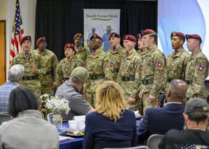 A group of soldiers in camouflage uniforms and maroon berets sing in front of an audience. A banner reads Small Farms Week in the background. The audience is seated with plates and drinks on the tables. An American flag is visible on the left.