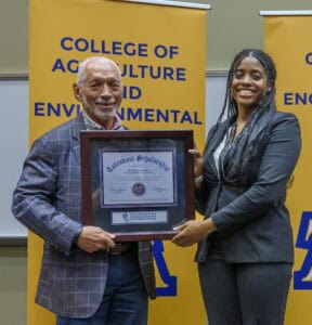 A smiling man and woman hold a framed Astronaut Scholarship certificate. They stand in front of yellow banners reading College of Agriculture and Environmental.