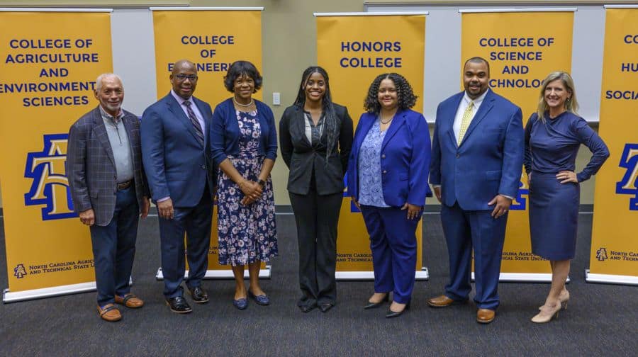 Seven professionally dressed people stand smiling in front of banners for different colleges at North Carolina A&T State University, including Agriculture and Environmental Sciences, Engineering, Honors College, and Science and Technology.