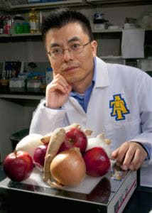 A man in a white lab coat with an A T logo sits at a lab table, resting his chin on his hand. In front of him are apples, onions, and ginger. Lab equipment and supplies are visible in the background.