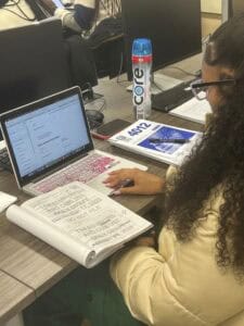 A person with long curly hair works at a desk with a laptop, notebook, and water bottle. The laptop displays a Google search, and the notebook has handwritten notes. A printed paper with 40012 is also visible.