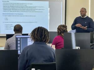 A man stands in front of a classroom facing students, presenting a slide with tax-related questions and information. Four students, seen from behind, sit at desks with computers and notebooks.