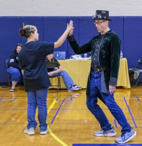 A man in a steampunk outfit and a young girl high-five in a gymnasium while people sit and watch in the background.