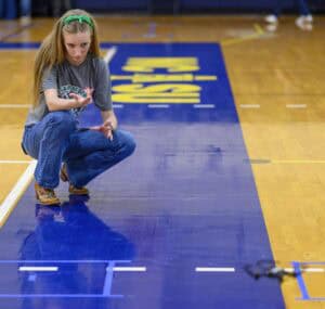 A girl crouches on a gym floor, focusing as she releases a small object, possibly a mechanical device, onto a marked blue strip on the court. She appears to be participating in a robotics or engineering activity.