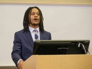 A man in a suit and tie with dreadlocks speaks at a podium in front of a whiteboard, appearing to give a presentation or lecture.