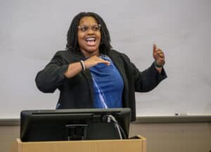 A person wearing glasses, a blue shirt, and a black blazer stands behind a podium, smiling and gesturing energetically with one hand raised, in front of a whiteboard.