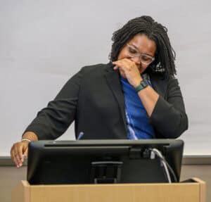 A person with glasses and shoulder-length hair stands at a podium, wearing a dark blazer and blue shirt. They are looking down with one hand covering their mouth, appearing thoughtful or emotional.