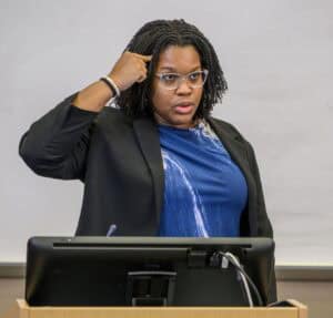 A woman with glasses and shoulder-length braids stands behind a podium, gesturing with her right hand near her temple as she speaks, wearing a blue top and black blazer in front of a whiteboard.