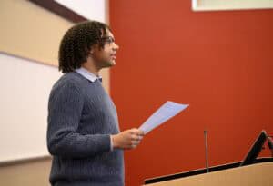 A person with curly hair and glasses stands at a podium, holding a sheet of paper and speaking in a classroom with a red wall and a whiteboard in the background.