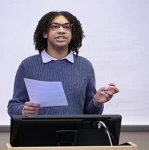 A person with curly hair and glasses stands at a podium, holding papers and speaking, with a whiteboard in the background.