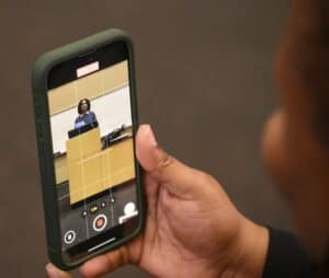 A person holds a smartphone that is recording or photographing a woman standing at a podium in a classroom or lecture hall. The phone screen shows the woman speaking behind the podium.