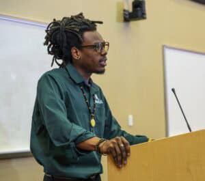 A person with dreadlocks, glasses, and a green shirt stands at a podium, speaking in a classroom or lecture hall setting. Whiteboards and a wall-mounted camera are visible in the background.
