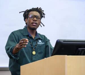 A person with dreadlocks wearing glasses and a green MANRRS shirt speaks at a podium, gesturing with one hand in front of a computer monitor and a white background.