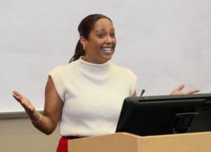A woman stands behind a podium, smiling and gesturing with her hands as she speaks, in front of a whiteboard in a classroom or lecture hall.